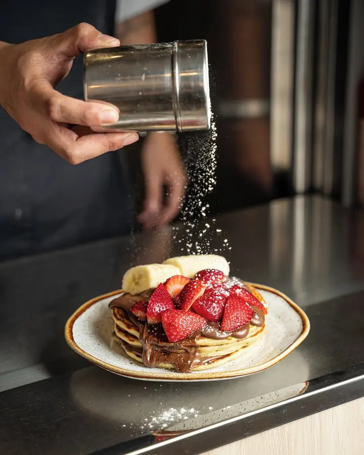 Mão a polvilhar açúcar sobre panquecas com Nutella, morangos e banana