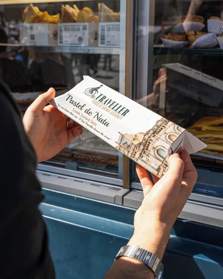 Mão a segurar uma caixa de pastéis de nata para levar, junto à montra